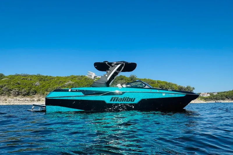 Slide: The Image of 2021 Malibu M240 boat on water, vibrant blue, clear sky, lush shoreline background. - 6
