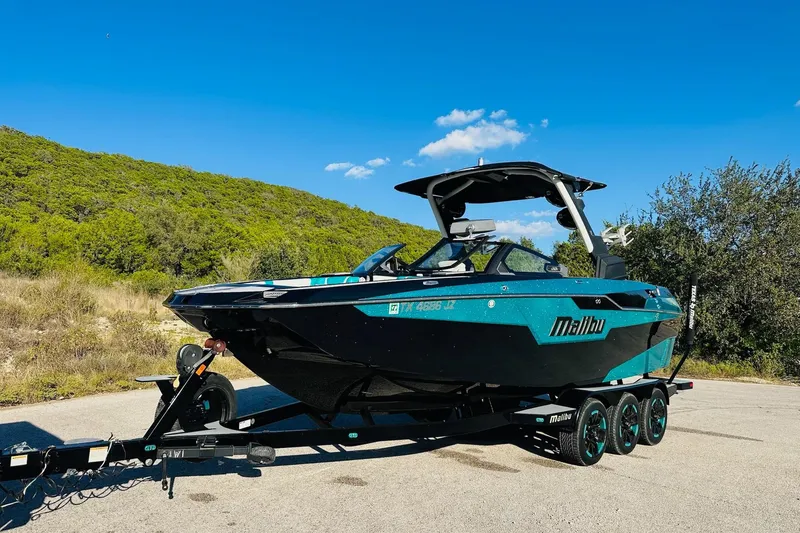 Slide: The Image of 2021 Malibu M240 boat on trailer, parked outdoors under clear blue sky. - 8