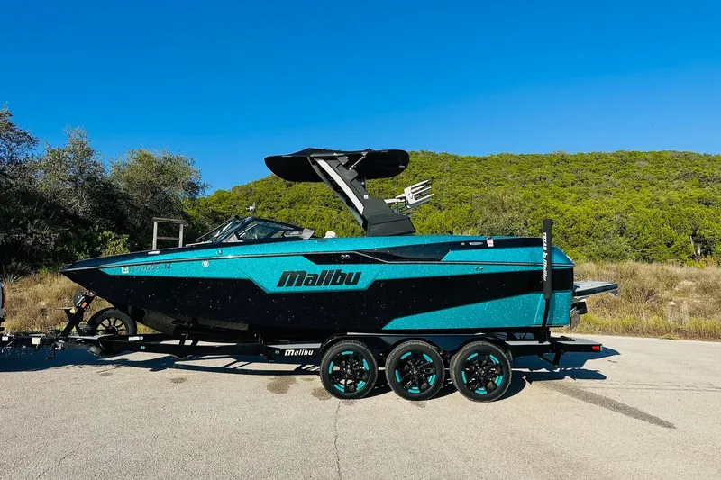 Slide: The Image of 2021 Malibu M240 boat on trailer, vibrant blue, parked outdoors with trees in background. - 9