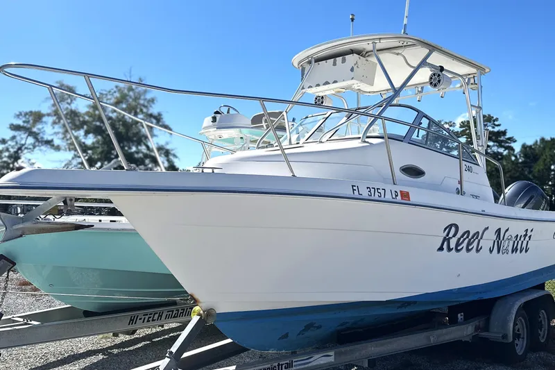The Image of 2000 Cobia 240 Walkaround boat on trailer, named "Reel Nauti," under clear blue sky. - 0