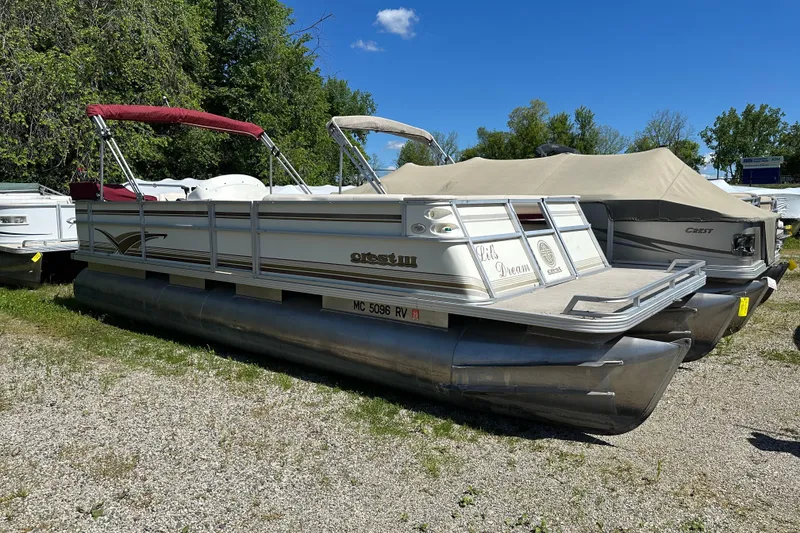 The Image of 1999 Crest 25 III Classic pontoon boat parked on gravel, surrounded by trees and other boats. - 1
