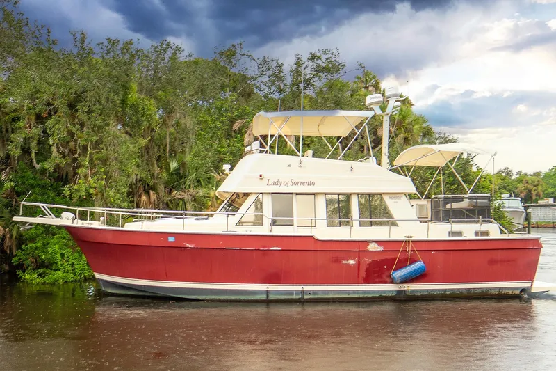 The Image of 2006 Mainship 43 Trawler, red hull, docked by lush greenery under cloudy sky. - 0