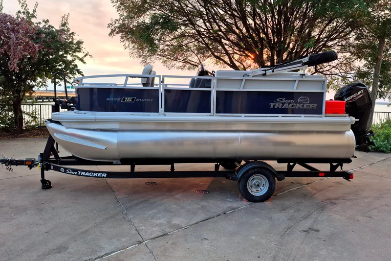 The Image of 2026 Sun Tracker Bass Buggy 16 XL Select pontoon boat on trailer, parked outdoors. - 0
