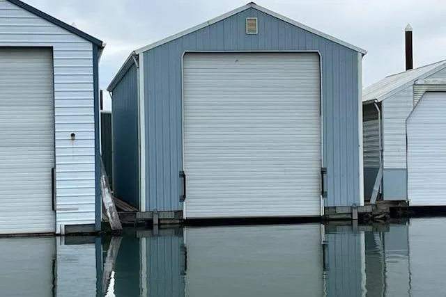 Slide: The Image of Custom 1980 boathouse with blue exterior and white garage door, reflected in calm water. - 3